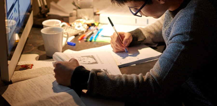 A young man sits at his desk studying with books, paper and a computer.