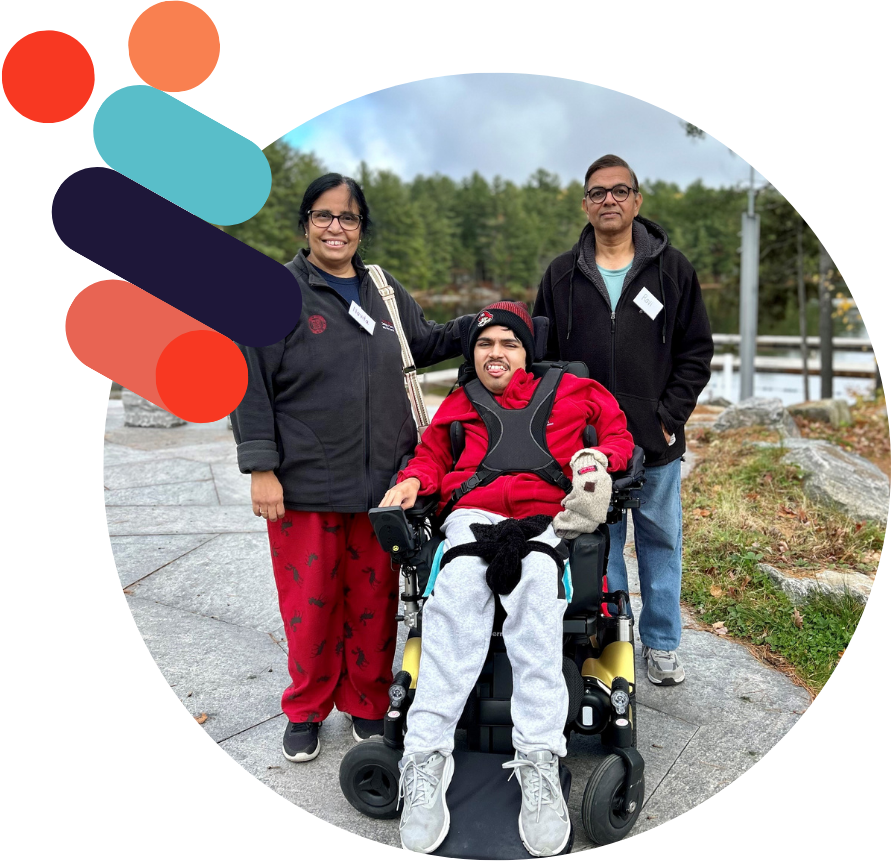 A person in a motorized wheelchair, dressed warmly in a red jacket, grey pants, and black and red toque, is smiling between two adults standing beside them. They are outdoors on a stone path, surrounded by trees and a lake in the background.