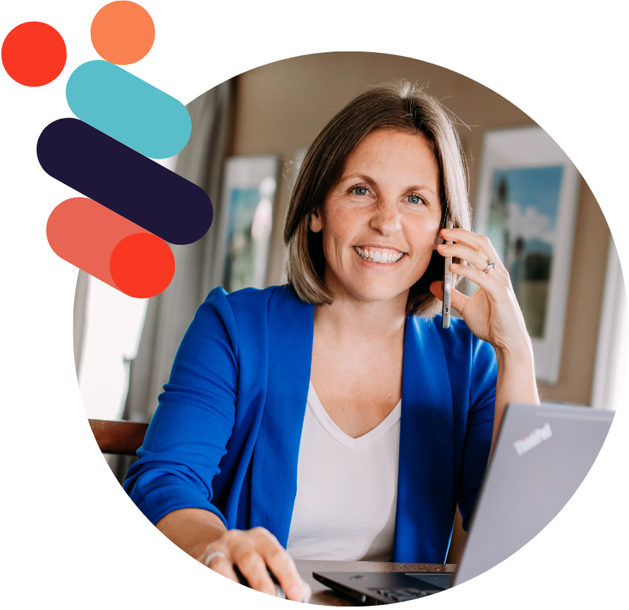 A smiling woman in a blue blazer talks on a cellphone while working on a laptop at a desk in a bright room with framed pictures on the wall.