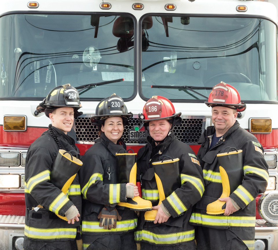 Four smiling firefighters in full turnout gear pose together in front of a white and red fire truck. Three of them are holding yellow and black rubber boots, symbolizing a "Fill the Boot" fundraising campaign.