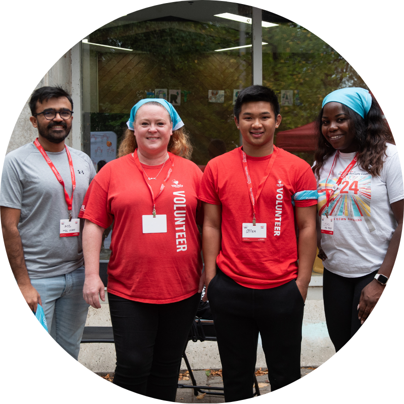 Quatre bénévoles devant un bâtiment avec badges et cordons rouges, portant des t-shirts rouges, gris et blanc.