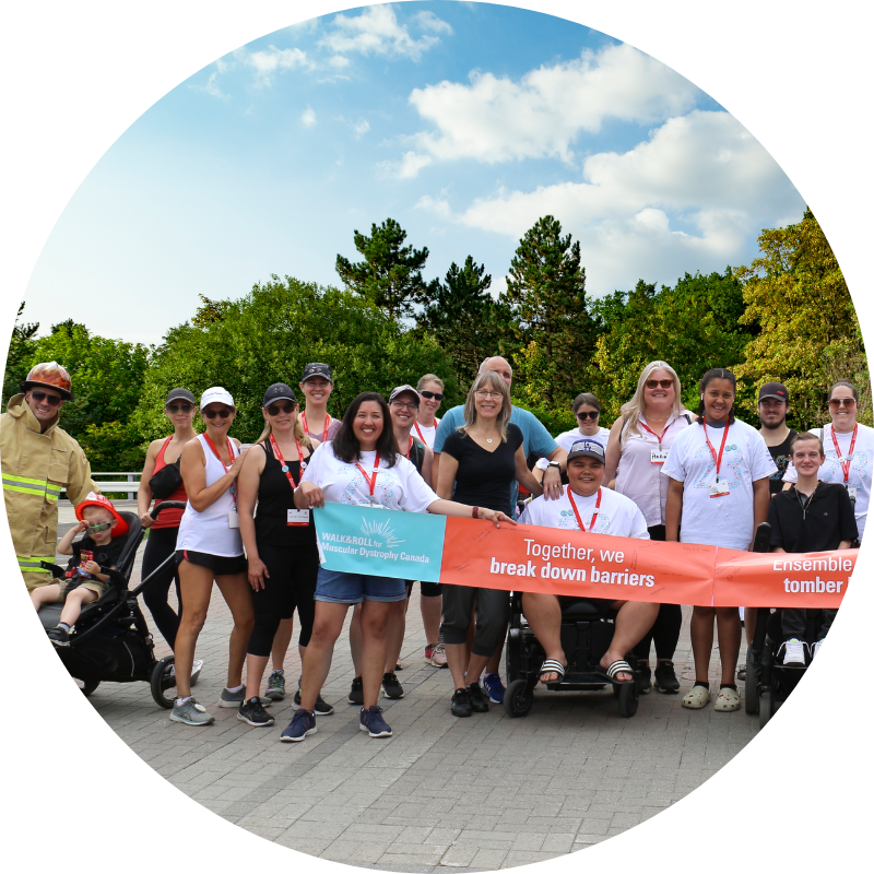 A diverse group of people, including individuals in wheelchairs, two firefighters in full gear, and others dressed casually, stand outdoors holding a large orange and blue banner that reads “Together, we break down barriers / ensemble, nous faisons tomber les barrières.” Trees and a partly cloudy sky are visible in the background.
