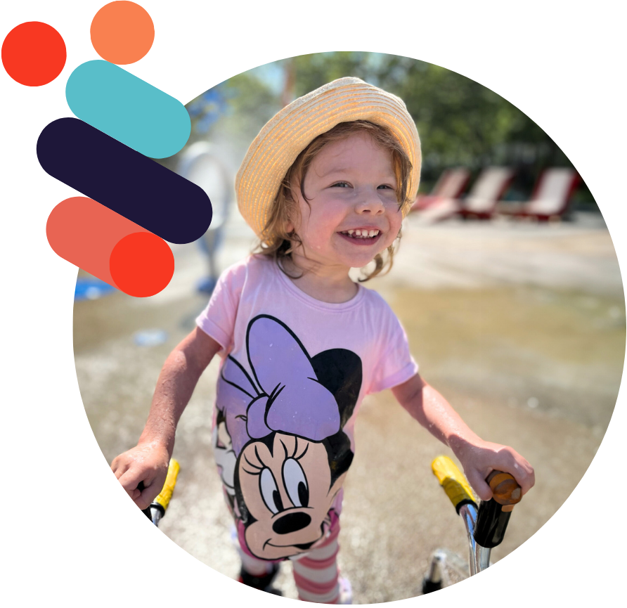 Jeune enfant souriante portant un chapeau de paille et un t-shirt rose joue, tenant les poignées de son aide à la mobilité avec de l’eau vaporisée en arrière-plan.