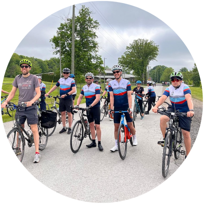 A group of cyclists wearing helmets and colorful gear ride together on a tree-lined street near a large building. The weather is cloudy but bright.