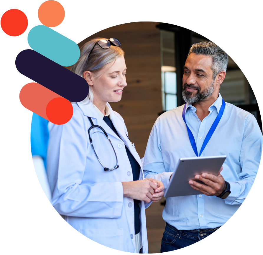 A group of healthcare professionals, including a doctor in a white coat and three nurses in blue scrubs, stand together while a man in a light blue shirt shows them information on a tablet. They appear engaged in discussion inside a modern medical facility.