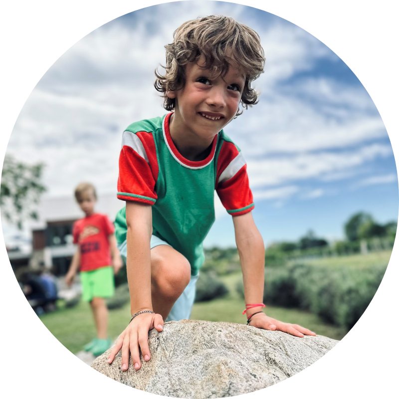Un jeune garçon aux cheveux bruns ondulés, portant un t-shirt vert et rouge, grimpe sur un grand rocher. Un autre enfant en t-shirt rouge et short vert se tient flou en arrière-plan.