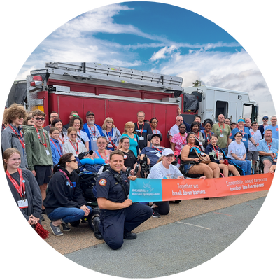 A large, diverse group of people, including firefighters and community members, pose outdoors in front of a red fire truck. They hold a long orange and blue banner and smile under a partly cloudy blue sky.