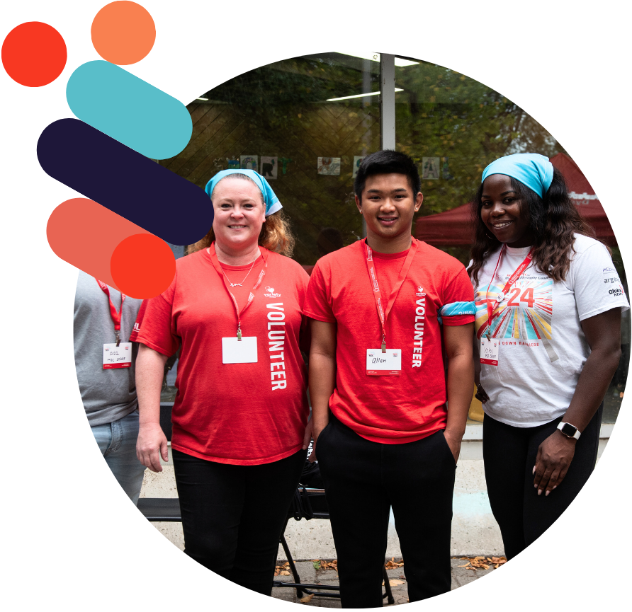 Three volunteers stand smiling outside a building, wearing volunteer badges and name tags on red lanyards.