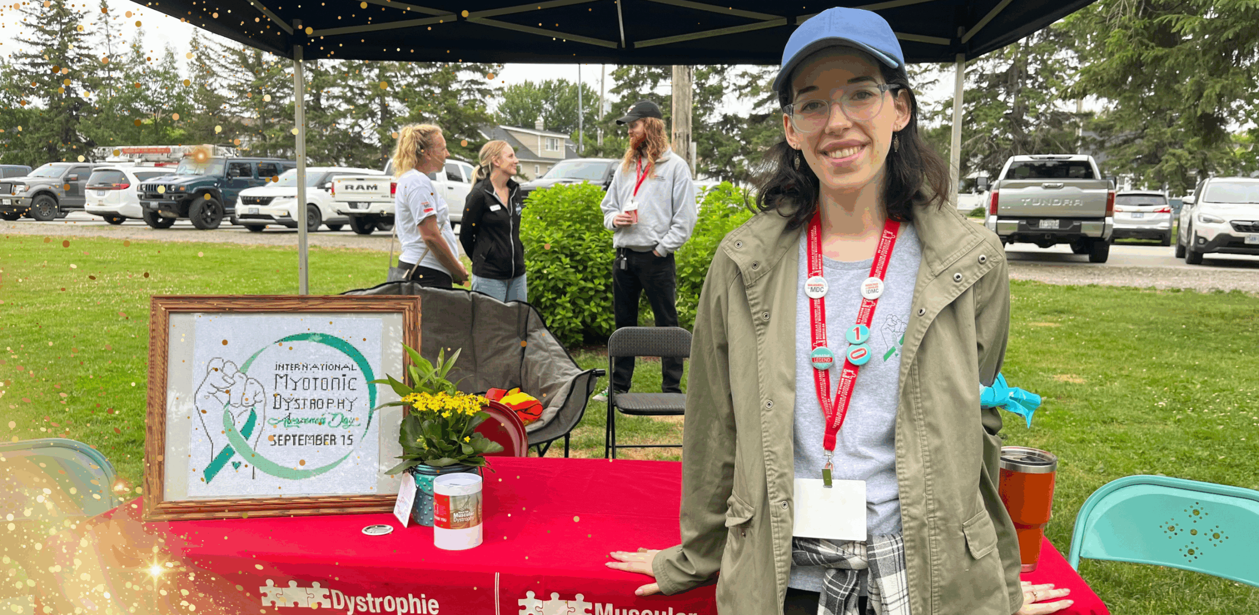 A smiling woman wearing glasses, a blue cap, and a red lanyard stands under a canopy at an outdoor event. In front of her is a table covered with a red cloth featuring muscular dystrophy organization logos, along with a framed sign promoting “International Myotonic Dystrophy Awareness Day – September 15,” a donation container, and a small bouquet of yellow flowers. Behind her, three people chat near parked cars and green shrubs. Gold sparkle effects are overlaid on the image.