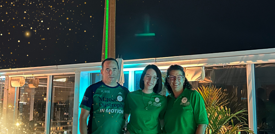 Three people standing together on a rooftop at night, smiling at the camera. They are wearing green Muscular Dystrophy Canada event shirts. Behind them are city buildings and the illuminated CN Tower glowing green. Decorative golden light effects appear in the sky above.