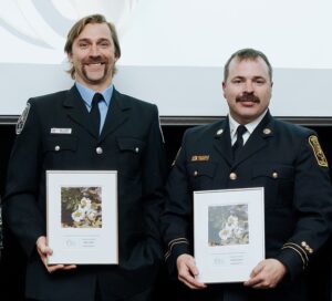 Portrait of two male firefighters dressed in formal attire holding a framed photo award. Both men have mustaches and dark hair.