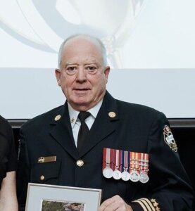 Portrait of an elderly male firefighter dressed in formal attire holding a framed photo award.
