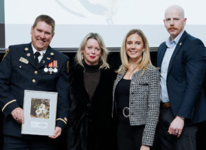 A group of people stand side by side, dressed in formal attire. The group includes 2 men and 2 women, with one name, a fire fighter in formal attire holding a framed photo award.