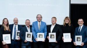 A group of people stand side by side, holding framed photo awards. The group is nicely dressed in formal attire and includes 2 women and 5 men.