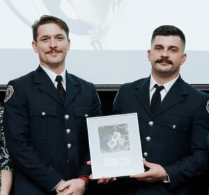 Portrait of two male firefighters dressed in formal attire holding a framed photo award. Both men have mustaches and dark hair.