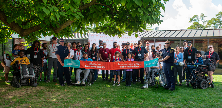 A diverse group of participants, holds a bilingual advocacy banner under a tree at a Walk and Roll for Muscular Dystrophy Canada event.