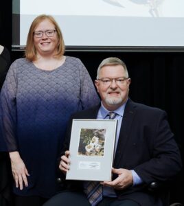 Portrait of a man in a wheelchair with glasses, a grey goatee and hair and a woman dressed in purple with glasses and red hair. The man is holding a framed photo award.