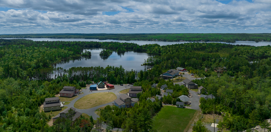 Aerial view of a lakeside community surrounded by dense green forest under a partly cloudy sky. Several houses and buildings are arranged along winding roads, with a central open grassy area. Multiple lakes and small islands are visible in the background, creating a scenic and peaceful natural setting.
